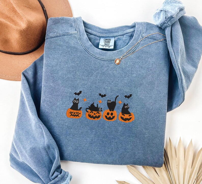 May include: A blue crewneck sweatshirt featuring an embroidered Halloween design. The design includes four black cats sitting on orange pumpkins with bats flying above. A gold necklace is draped on the sweatshirt.
