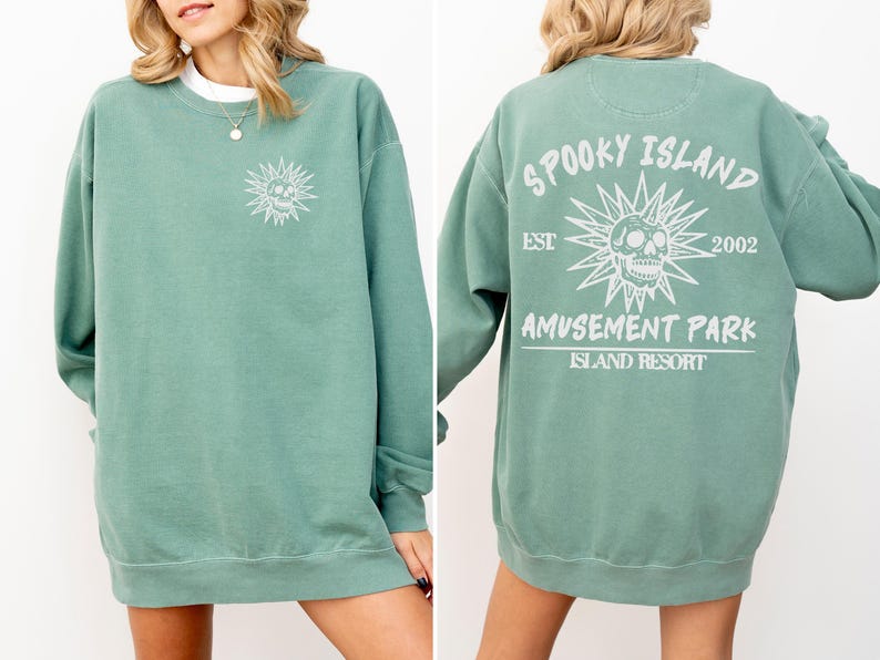 May include: A seafoam green sweatshirt with a crew neck. The front features a white graphic of a skull within a sunburst design. The back displays the text "Spooky Island Amusement Park Island Resort" with the year "2002" and a similar skull graphic.
