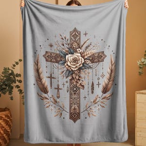 May include: A light gray blanket featuring a detailed brown cross design. The cross is adorned with floral and feather accents, with a central rose and smaller crosses. The design is symmetrical and decorative.
