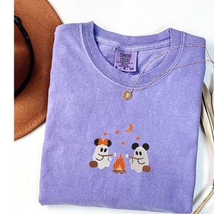 May include: A purple short-sleeve t-shirt featuring an embroidered graphic of two ghosts roasting marshmallows over a campfire. The ghosts wear mouse-ear headbands. The shirt has a Comfort Colors label at the neck and a crew neckline.