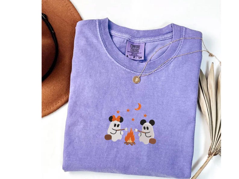 May include: A purple short-sleeve t-shirt featuring an embroidered graphic of two ghosts roasting marshmallows over a campfire. The ghosts wear mouse-ear headbands. The shirt has a Comfort Colors label at the neck and a crew neckline.