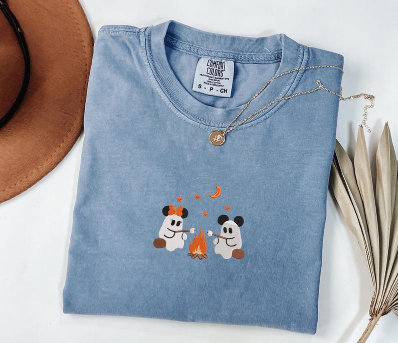 May include: A blue t-shirt with a Comfort Colors label in the collar, featuring an embroidered graphic of two ghosts with mouse ears roasting marshmallows over a campfire, surrounded by orange stars and a crescent moon on the chest area.