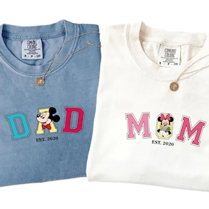 May include: A blue t-shirt with the text DAD and a Mickey Mouse graphic, and a white t-shirt with the text MOM and a Minnie Mouse graphic. Both shirts feature the text EST. 2020 below the graphics and have Comfort Colors brand labels at the neck.