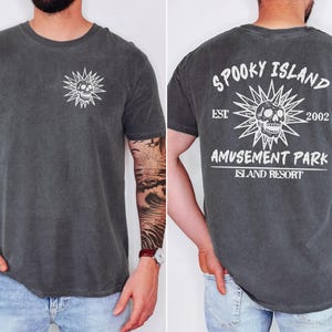 May include: Dark gray t-shirt with a white skull and sunburst graphic on the front and back. The back of the shirt reads "Spooky Island Amusement Park Island Resort" with the year "2002".