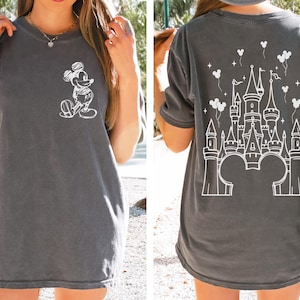 May include: A charcoal gray short-sleeve t-shirt featuring a white line-art graphic of Mickey Mouse on the left chest. The back of the shirt displays a large white line-art illustration of a castle with Mickey-shaped balloons and stars.