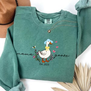 May include: Teal-colored sweatshirt with a Mama Goose graphic. The design features a cartoon goose wearing a blue hat and yellow bow, surrounded by flowers, butterflies, and a bee. The text "mama goose" and "Est. 2025" are also included.