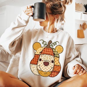 May include: Cream-colored sweatshirt featuring a patchwork pumpkin design with a smiling face and bear ears. The pumpkin is made of various patterned fabrics in fall colors. A black mug is held up to the face.