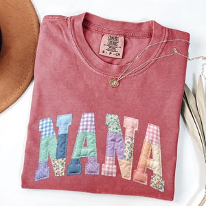 May include: A red Comfort Colors t-shirt with the word "NANA" in patchwork letters. The letters are made of various patterned fabrics, including gingham and floral prints. A gold necklace with a pendant rests on the shirt.