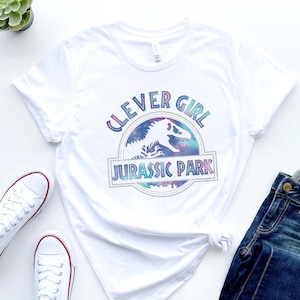 May include: White t-shirt featuring a colorful graphic with the text "CLEVER GIRL" above a dinosaur silhouette and the words "JURASSIC PARK". The shirt is paired with blue jeans and white sneakers.