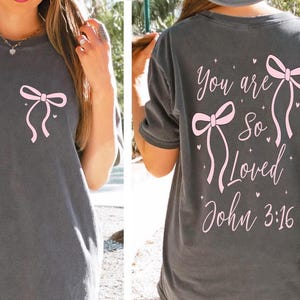 May include: Dark gray t-shirt with a pink bow graphic on the front and back. The back of the shirt features the text "You are So Loved John 3:16" in pink script. The shirt has a casual, relaxed fit.