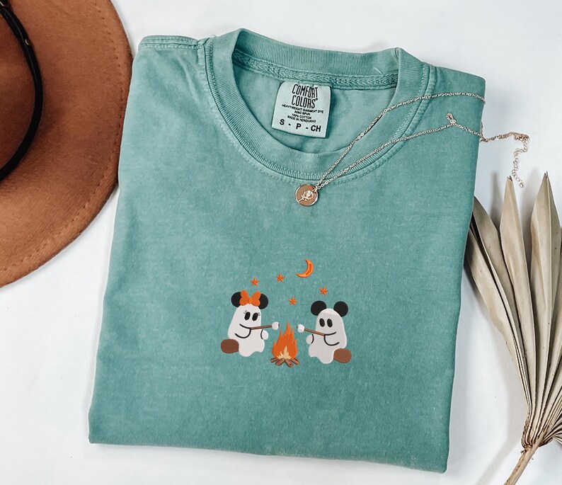 May include: A sage green short-sleeve t-shirt featuring an embroidered graphic of two ghosts with mouse ears roasting marshmallows over a campfire under a crescent moon and stars. The shirt has a Comfort Colors brand label in the neck.