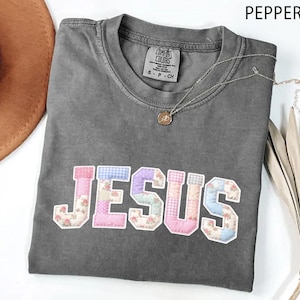 May include: A gray t-shirt with the word "JESUS" appliquéd across the front in a patchwork design of floral, gingham, and solid fabrics. A silver necklace with a pendant and a brown hat are also visible.