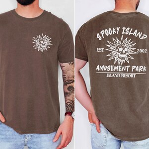 May include: Brown t-shirt with a white skull and sunburst graphic on the front and back. The back of the shirt reads "Spooky Island Amusement Park Island Resort" with "Est 2002".