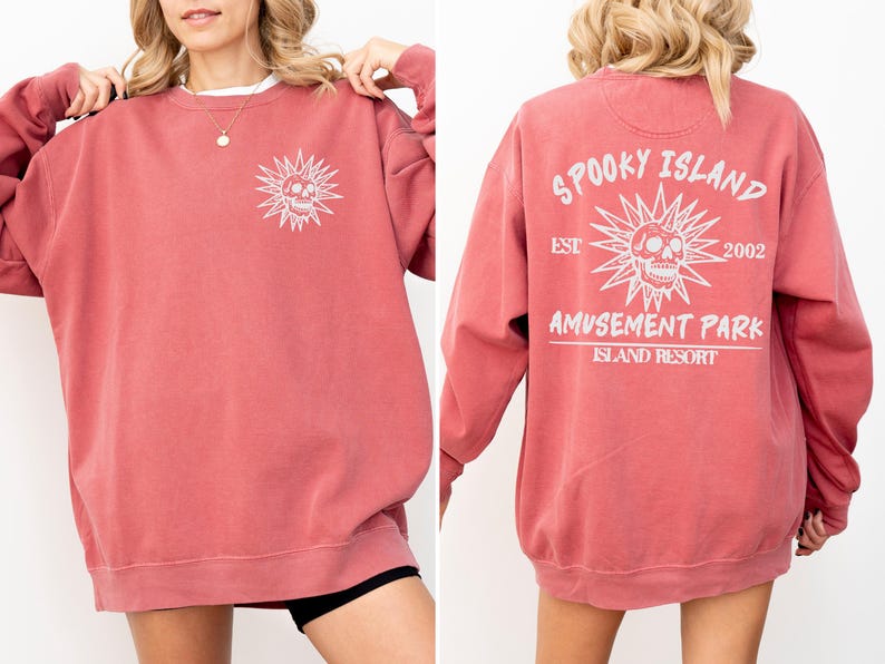 May include: A dusty rose-colored sweatshirt with a white graphic of a skull within a sunburst design on the front. The back features the text "Spooky Island Amusement Park Island Resort" with a skull graphic and the year "2002".
