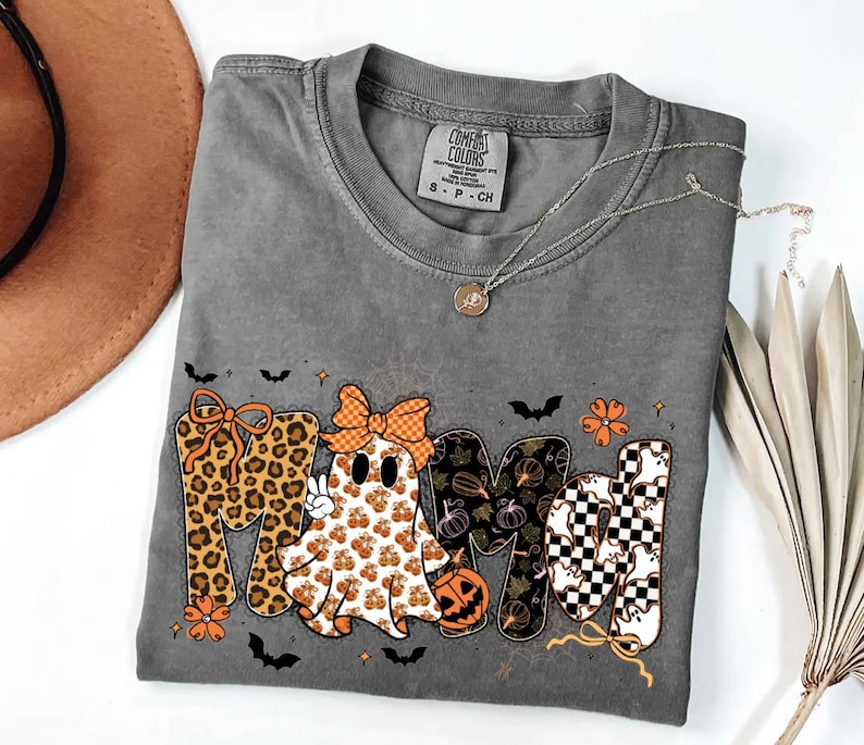 May include: A charcoal gray t-shirt featuring a graphic that spells MAMA, where the first letter is leopard print, the second is a ghost with a bow, the third has a pumpkin pattern, and the fourth is a black and white checkerboard with ghosts.
