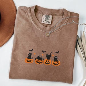 May include: Brown Comfort Colors t-shirt with a crew neck and short sleeves. The shirt features an embroidered design of four black cats sitting in orange jack-o'-lanterns with bats above them. A silver necklace with a pendant is draped across the shirt.