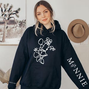 May include: Black hooded sweatshirt featuring a white line drawing of Minnie Mouse on the front. The right sleeve has the word "MINNIE" in white, with a floral design. The text "SINCE 1928" is printed on the left sleeve.