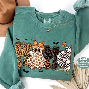 May include: A light green crewneck sweatshirt featuring a graphic print that spells MAMA. The letters incorporate leopard print, a ghost wearing an orange bow, pumpkin patterns, and a black and white checkerboard pattern with small ghost icons.