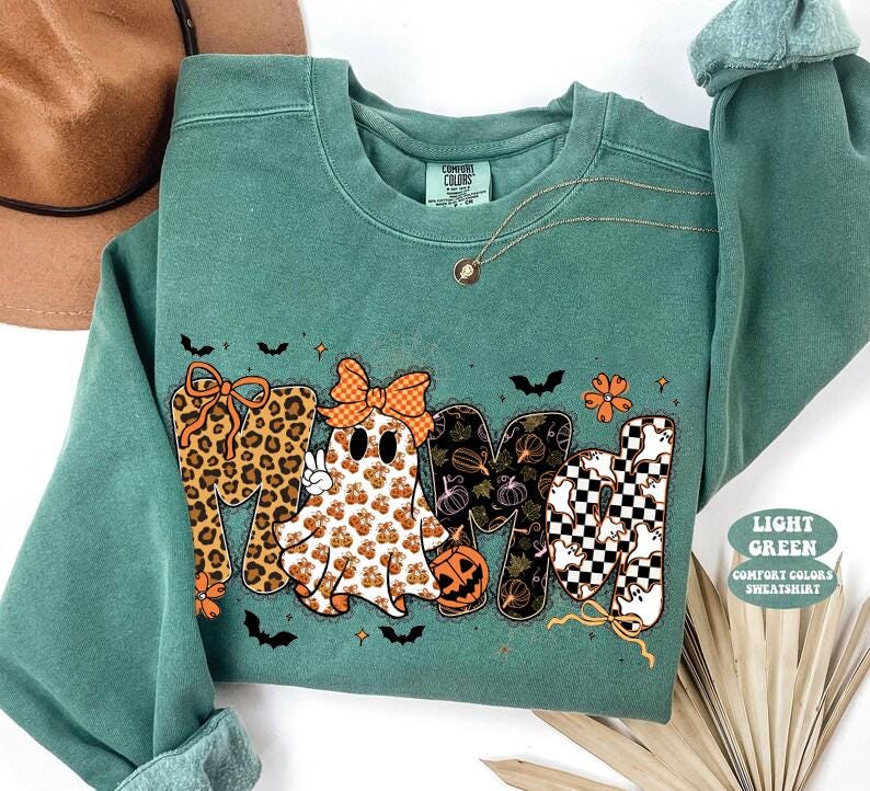 May include: A light green crewneck sweatshirt featuring a graphic print that spells MAMA. The letters incorporate leopard print, a ghost wearing an orange bow, pumpkin patterns, and a black and white checkerboard pattern with small ghost icons.