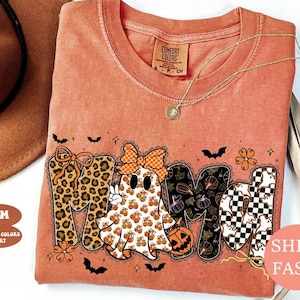 May include: A burnt orange t-shirt featuring a graphic print that spells MAMA. The letters are decorated with leopard print, a ghost wearing a checkered bow, pumpkin patterns, and a black and white checkerboard pattern with small ghosts.