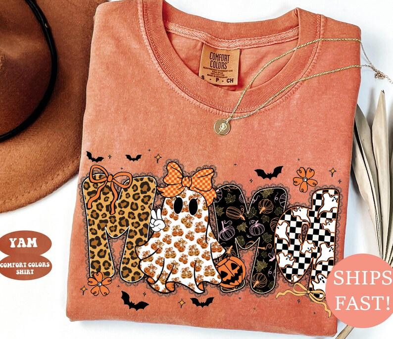 May include: A burnt orange t-shirt featuring a graphic print that spells MAMA. The letters are decorated with leopard print, a ghost wearing a checkered bow, pumpkin patterns, and a black and white checkerboard pattern with small ghosts.