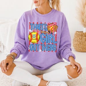 May include: A lilac-colored sweatshirt with the text "That's My Girl Out There" in blue and red. The design includes a softball, bat, and glove. The softball has the number 15 and the name "Amelia" on it.