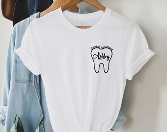 May include: White t-shirt with rolled-up sleeves. The shirt features a black graphic of a tooth with the text "Dental Assistant Ashley" above the tooth. The shirt is hanging on a wooden hanger.