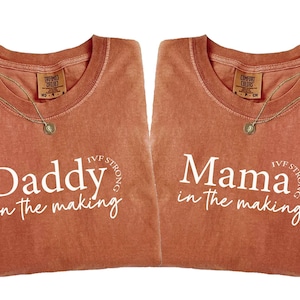 May include: Two terracotta-colored t-shirts with the phrases "Daddy in the making" and "Mama in the making" in white script. Each shirt has a gold necklace with a pendant. The shirts are from the "Comfort Colors" brand.