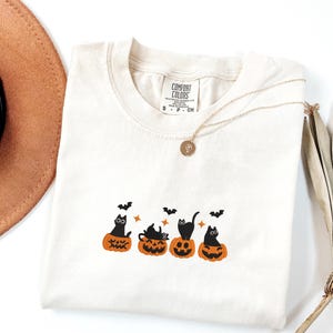 May include: Cream-colored Comfort Colors t-shirt featuring a Halloween design. The design includes black cats sitting in carved orange pumpkins, with bats flying above. A gold necklace with a pendant is draped across the shirt.