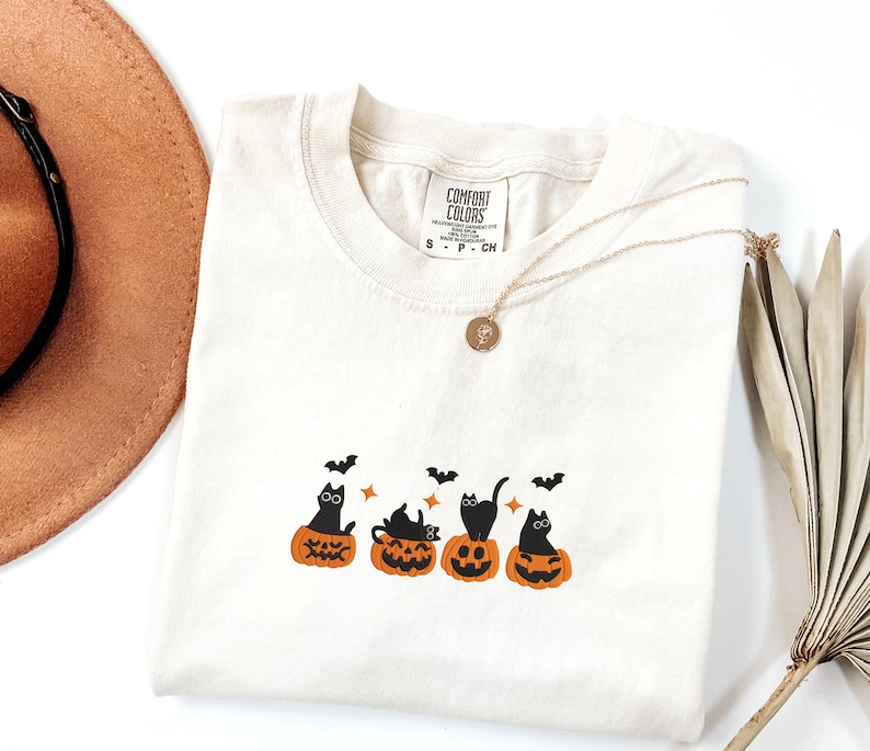 May include: Cream-colored Comfort Colors t-shirt featuring a Halloween design. The design includes black cats sitting in carved orange pumpkins, with bats flying above. A gold necklace with a pendant is draped across the shirt.