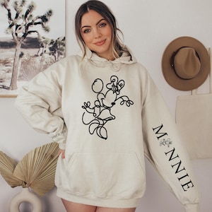 May include: Cream-colored hoodie featuring a black line drawing of Minnie Mouse on the front. The right sleeve has the word "MINNIE" in black, with a small floral design. The hoodie has a front pocket and a drawstring hood. The text "SINCE 1928" is printed on the left sleeve.