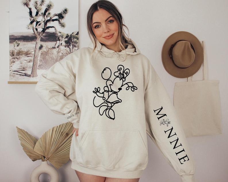 May include: Cream-colored hoodie featuring a black line drawing of Minnie Mouse on the front. The right sleeve has the word "MINNIE" in black, with a small floral design. The hoodie has a front pocket and a drawstring hood. The text "SINCE 1928" is printed on the left sleeve.