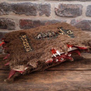 May include: A handcrafted, brown, furry book with the word "ROZZEN" in gold lettering. The book has a red, toothy maw design on the edges, with white teeth and red accents. The book is set against a brick wall and wooden surface.