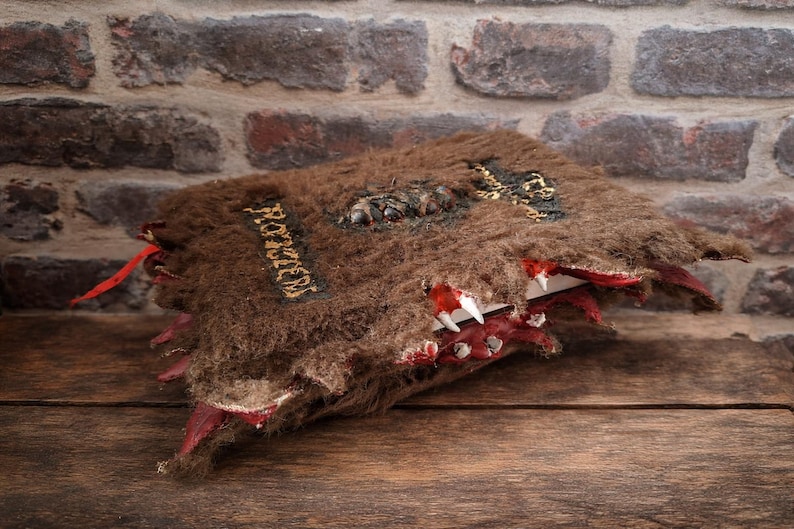 May include: A handcrafted, brown, furry book with the word "ROZZEN" in gold lettering. The book has a red, toothy maw design on the edges, with white teeth and red accents. The book is set against a brick wall and wooden surface.