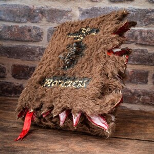 May include: A brown, furry book with the word "Monsters" in gold lettering. The book has a textured cover and red accents, with a red ribbon bookmark. The book's edges are jagged, with white and red details.