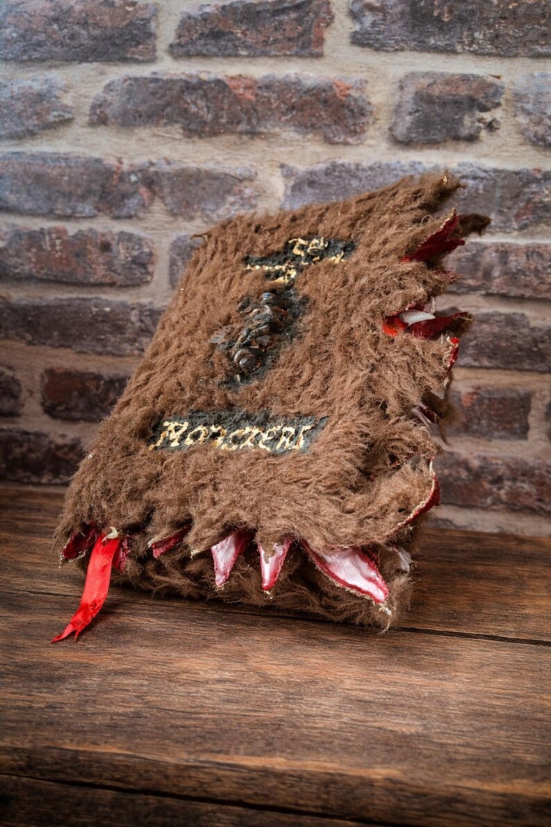 May include: A brown, furry book with the word "Monsters" in gold lettering. The book has a textured cover and red accents, with a red ribbon bookmark. The book's edges are jagged, with white and red details.
