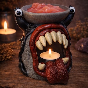 Może przedstawiać: Ceramiczny podgrzewacz do olejków o tematyce potworów z miseczką na woski zapachowe i miejscem na świeczkę typu tealight. Podgrzewacz ma czerwone usta z dużymi zębami, teksturowany język i dwa ruchome oczy. Miseczka zawiera różowe woski zapachowe.