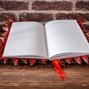 May include: An open notebook with lined pages rests on a red and brown decorative base with a dragon scale design. A red ribbon bookmark is visible. The background features a brick wall and a wooden surface.