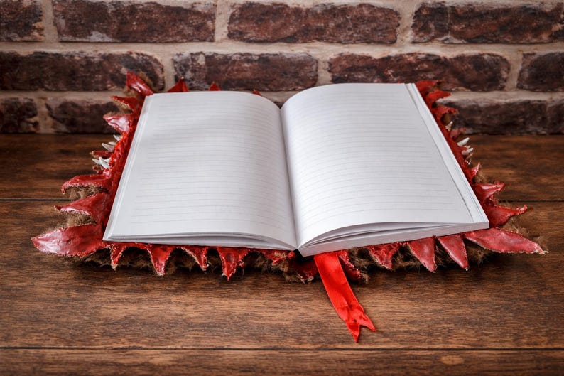 May include: An open notebook with lined pages rests on a red and brown decorative base with a dragon scale design. A red ribbon bookmark is visible. The background features a brick wall and a wooden surface.