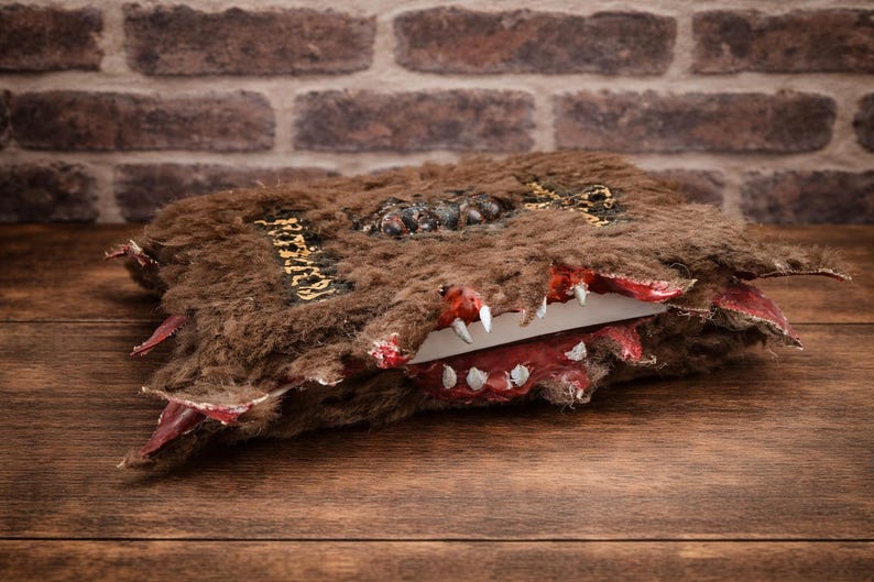 May include: A brown, furry book with a monster-like design. The book has a red, toothy maw and gold lettering on the spine. The book is open, revealing white pages. The background is a brick wall and a wooden surface.