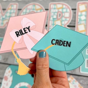 Puede incluir: Dos gorros de graduaci&oacute;n, uno rosa con un lazo y el nombre "RILEY" y otro azul verdoso con el nombre "CADEN".
