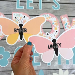 May include: Two pink and yellow butterfly-shaped paper cutouts with the names "Ryder" and "Linley" written on them. The butterflies are on a wooden background with a floral pattern.