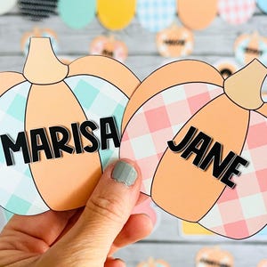 May include: Two orange pumpkin-shaped paper cutouts with gingham patterns. The pumpkin on the left has a blue and white gingham pattern and the name "MARISA" written in black. The pumpkin on the right has a pink and white gingham pattern and the name "JANE" written in black.