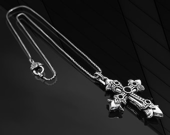 May include: A silver chain necklace with a large, ornate silver cross pendant featuring black gemstones. The cross has a detailed design with intricate flourishes.