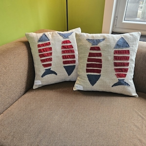 Peut inclure: Deux coussins carrés en tissu beige clair, chacun orné de deux motifs de poissons appliqués. Les poissons sont constitués de bandes horizontales de tissu bleu foncé et rouge profond, créant un motif rayé sur le fond clair.
