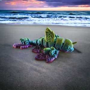 May include: A colorful, 3D printed, articulated, butterfly-like creature with iridescent wings and a green body. The creature is lying on a sandy beach.