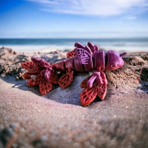 May include: A pink and red 3D printed sea creature with intricate details. The creature has a long tail and wings, and is made of a smooth, plastic-like material.