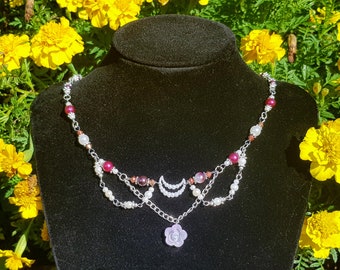 May include: A silver chain necklace with a pink flower pendant, white pearls, and burgundy beads. The necklace is displayed on a black mannequin against a backdrop of yellow flowers.