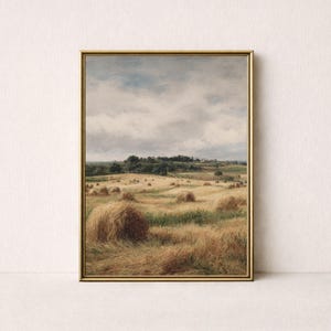 Op de afbeelding: Een ingelijst landschapsschilderij toont een veld met gouden hooibalen onder een bewolkte hemel. Het kunstwerk toont een landelijke scène met glooiende heuvels, bomen en een zacht, gedempt kleurenpalet, dat rust uitstraalt.