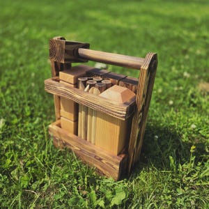 May include: A wooden toy set with a carrying handle. The set includes various geometric shapes, such as blocks, cylinders, and pyramids, all made of light-colored wood. The wooden carrier has a dark brown stain.